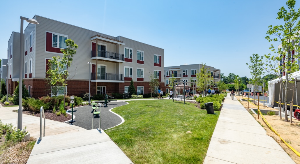 A modern apartment complex with two-story buildings, landscaped lawns, walking paths, young trees, lampposts, and outdoor seating areas on a sunny day. People are visible in the distance.