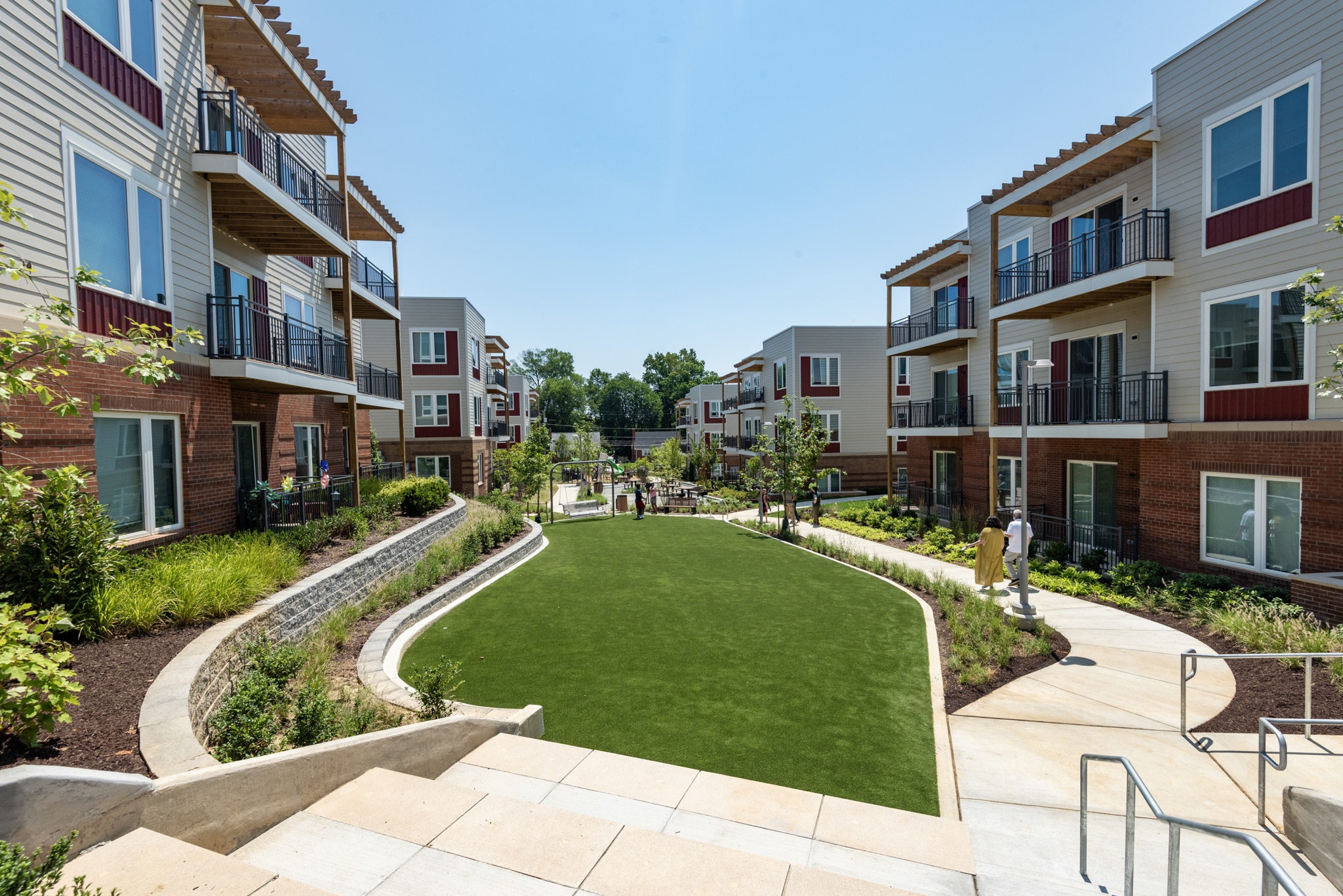 Modern apartment buildings with balconies surround a central green lawn courtyard, landscaped with plants and pathways, under a clear blue sky. A few people walk and relax in the outdoor communal space.