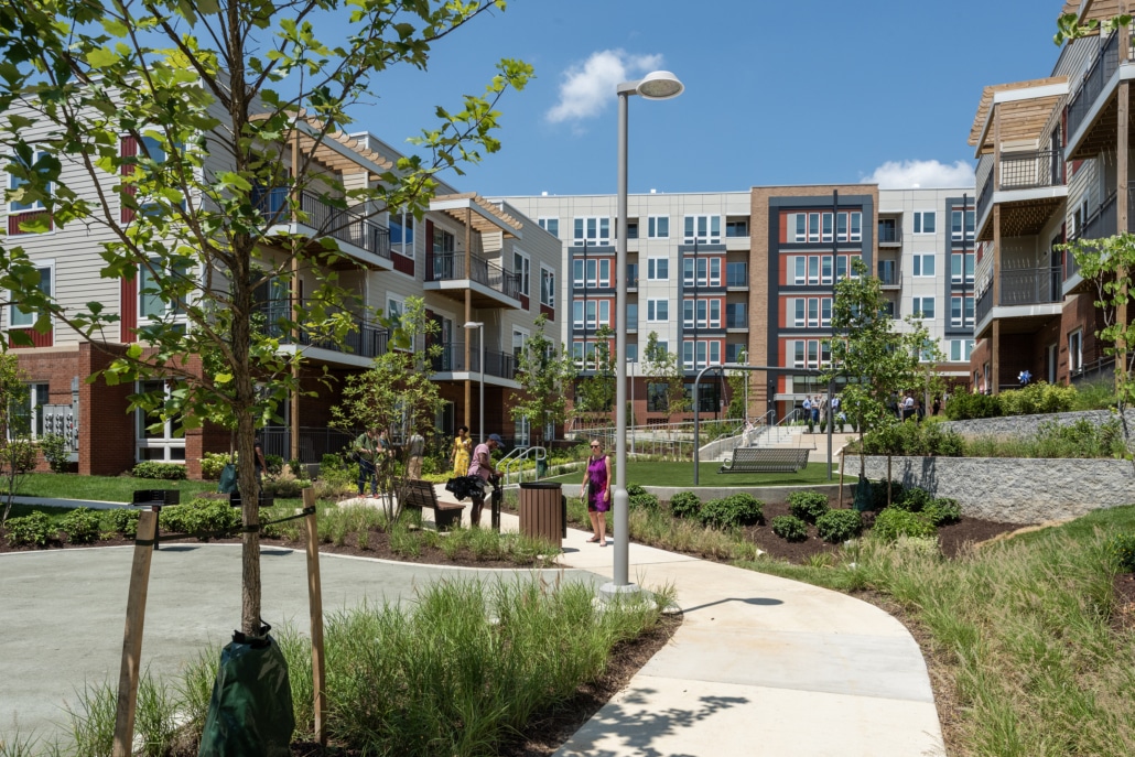 A landscaped courtyard in a modern apartment complex with people walking and relaxing on a sunny day. The buildings have balconies and large windows, and there are trees, paths, and benches throughout the area.