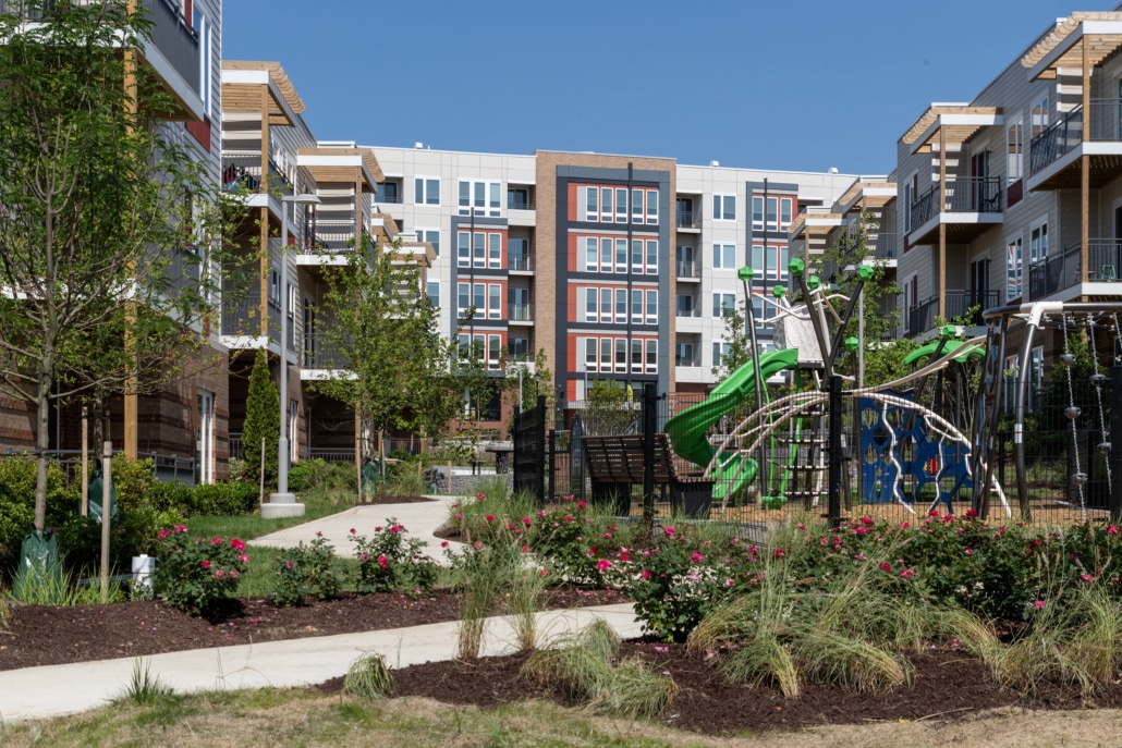 A modern apartment complex with multiple mid-rise buildings, landscaped gardens, and a children’s playground featuring slides and climbing equipment under a clear blue sky.