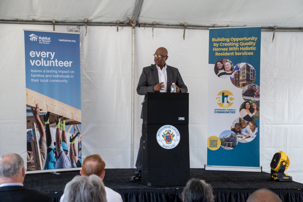 A man speaks at a podium under a white tent, with banners for Habitat for Humanity and affordable housing beside him. Audience members are seated and listening in the foreground.