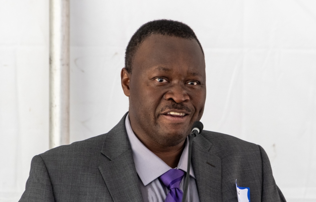 A man wearing a gray suit, lavender shirt, and purple striped tie stands at a podium with a microphone, speaking. He has short hair and a name tag on his jacket. A white background is behind him.