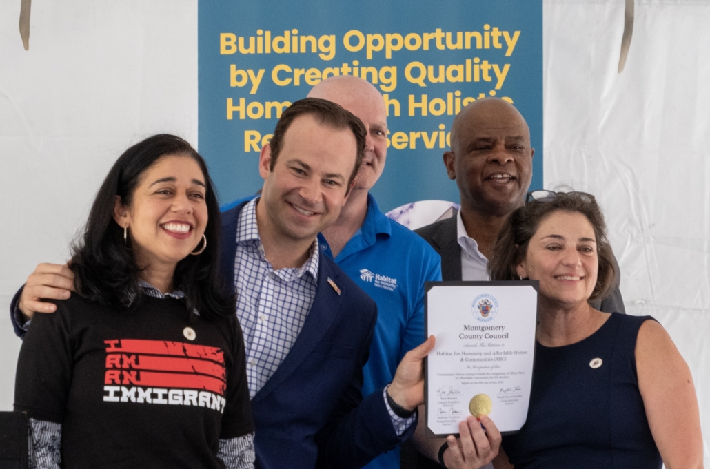 Group of five smiling people stand together; one woman holds up an official Montgomery County Council certificate. A sign behind them reads “Building Opportunity by Creating Quality Homes and Holistic Resident Services.”.