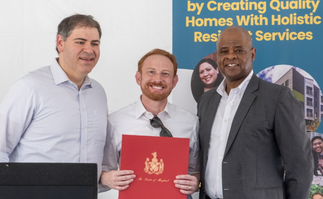 Three men in business attire stand together smiling; the man in the center holds a red folder with a gold crest. Behind them is a banner about creating quality homes with holistic residential services.