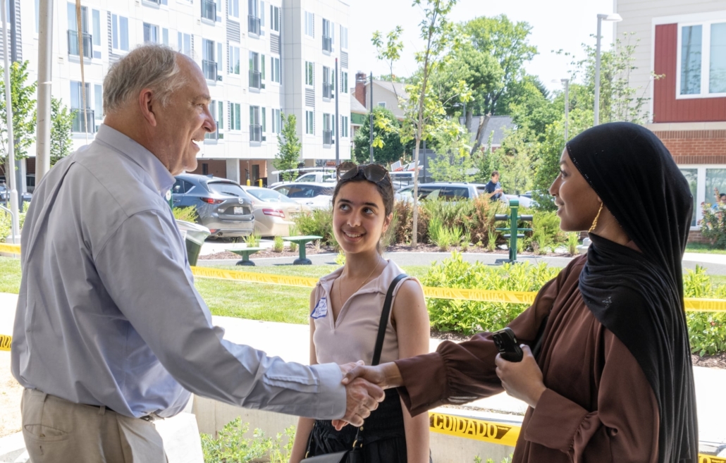 An older man in a light blue shirt shakes hands with a woman in a brown hijab outdoors, while a young woman standing between them smiles. Buildings and trees are in the background.