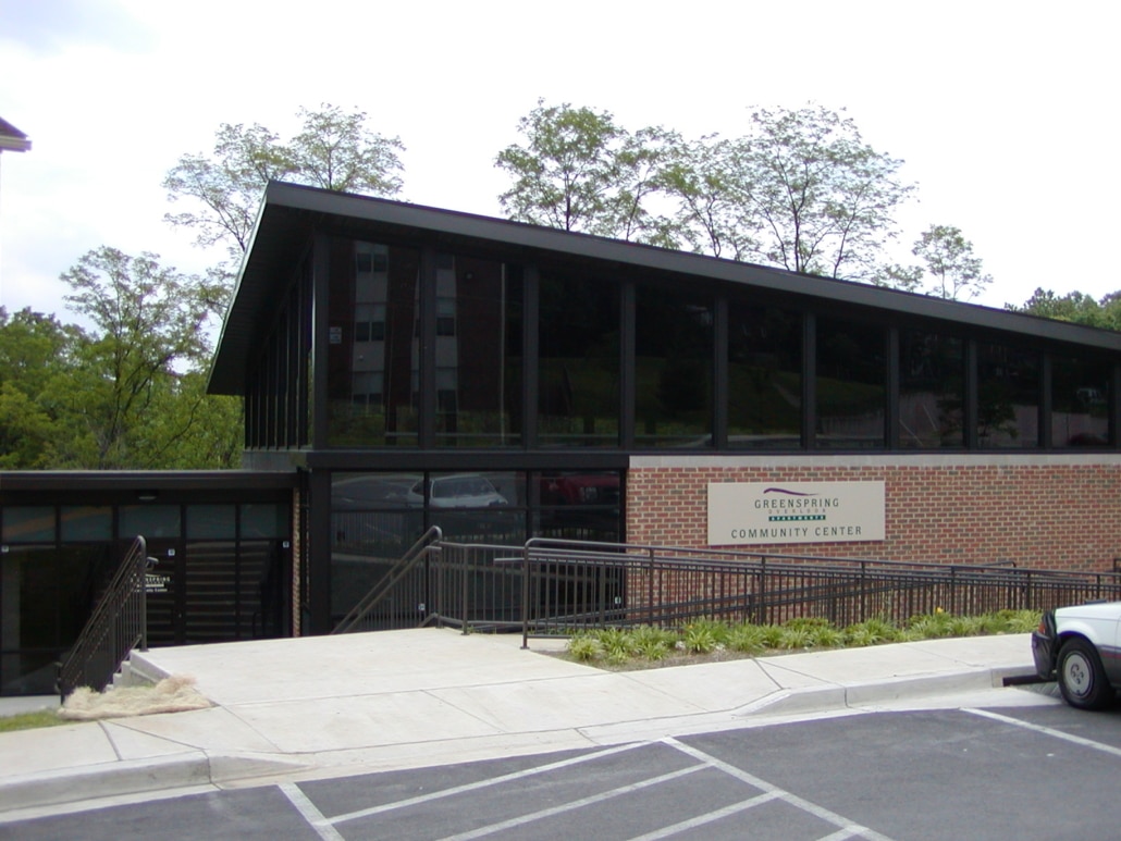 A modern brick and glass building with a slanted roof, labeled Greenspring Community Center, is pictured near a small parking lot with ramps, stairs, and surrounding greenery.
