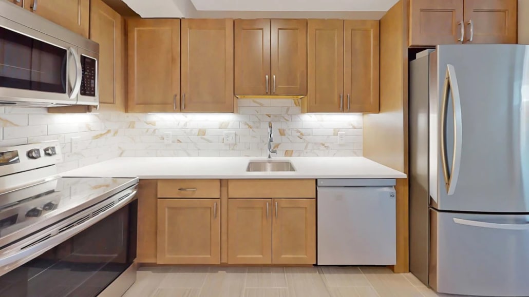 Modern kitchen with wood cabinets, stainless steel appliances including a stove, microwave, and refrigerator, white countertops, a built-in sink, a dishwasher, and a white marble-patterned backsplash.