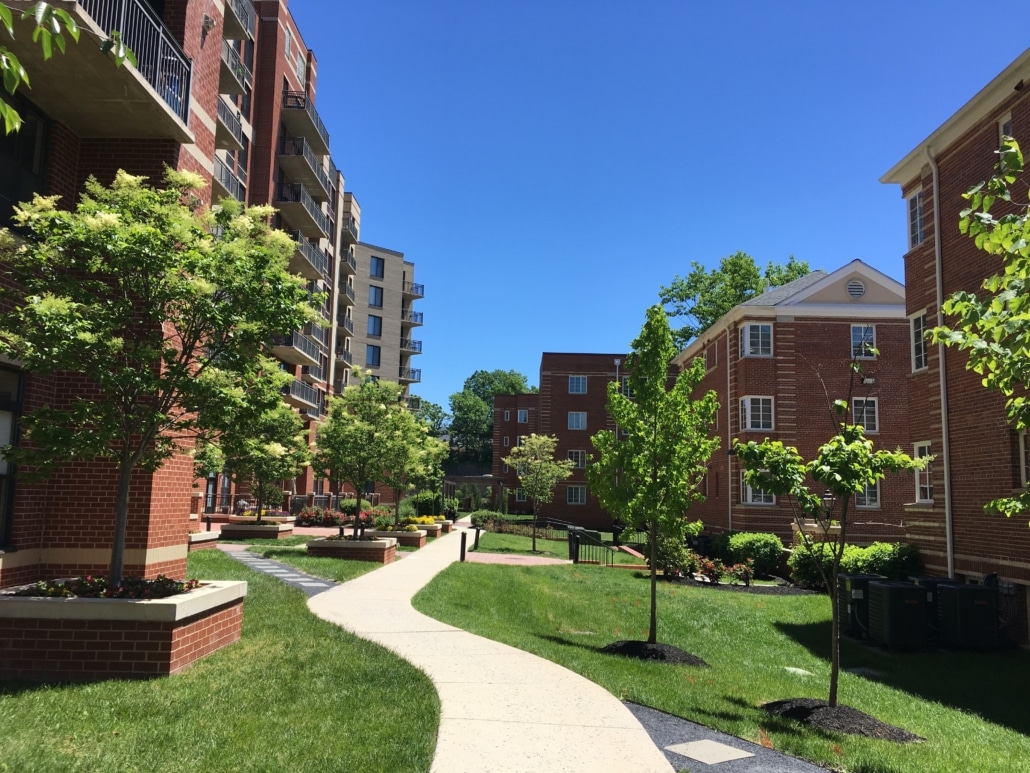 A sunny day in a residential complex with red brick apartment buildings, green lawns, young trees, benches, and a winding sidewalk, all under a clear blue sky.
